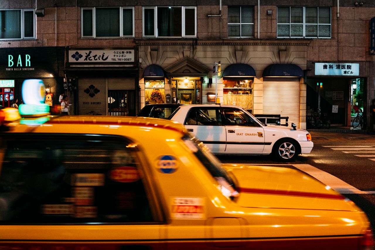 gallery-2 Dynamic Tokyo street scene featuring yellow taxis and illuminated storefronts at night.